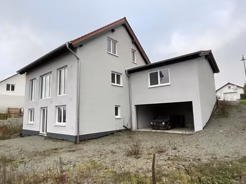 Zwangsversteigerung Einfamilienhaus in Nußbach – Titelfoto: Gartenseite mit verputzter Fassade, Garagenöffnung und geschottertem Gelände.