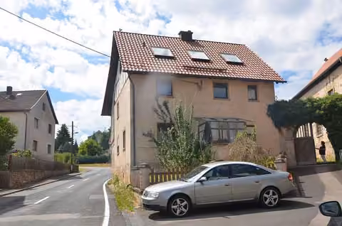 Zwangsversteigerung Zweifamilienhaus in Grabfeld, Wölfershausen – Straßenansicht des Wohnhauses mit Zwischenbau und davor parkendem Pkw.